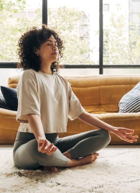 A woman with curly hair sits on a living room floor and meditates
