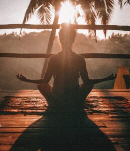 Woman sitting in meditative pose on wood deck at sunrise, stress reducing activity